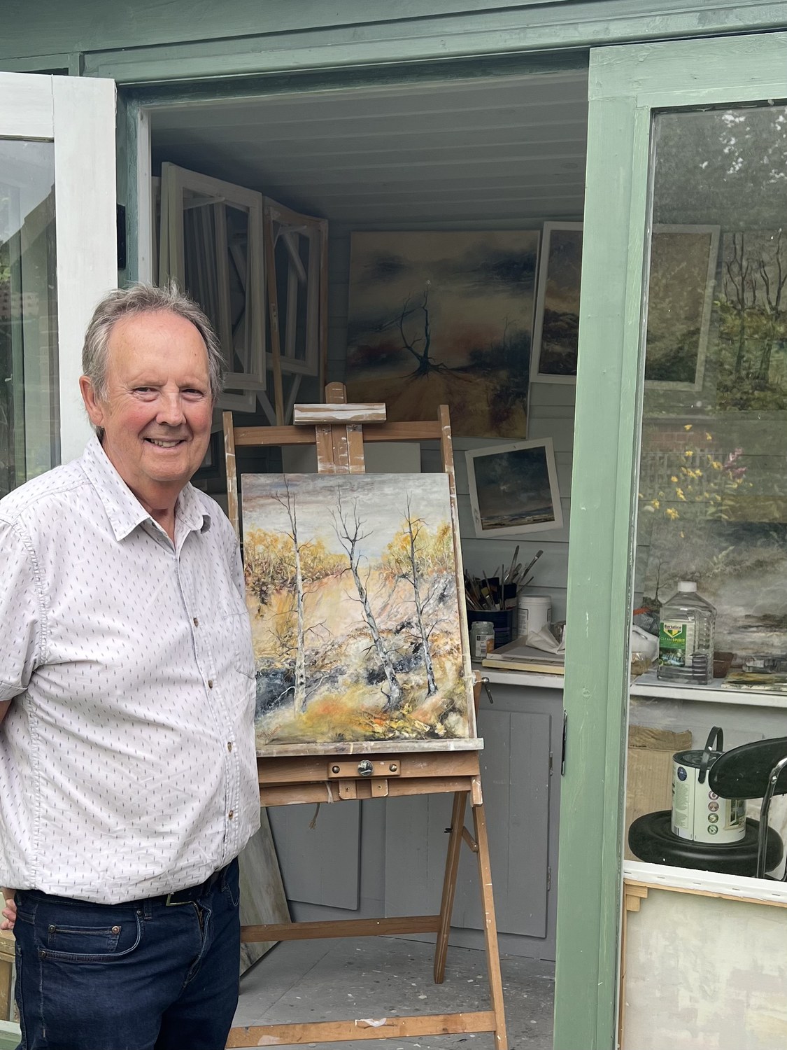Neil Williams at the door of his studio, a painting of birches on the easel behind him